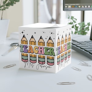 May include: A white cube-shaped notepad with a colourful illustration of pencils and the word "TEACHER" in a retro font. The notepad is personalised with the name "Mrs. Johnson".