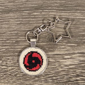 May include: A round, silver-toned keychain with a cross-stitch design. The design features a red and black spiral pattern on a white background. The keychain has a star-shaped clasp.