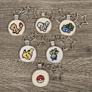 May include: Keychains featuring cross-stitched designs of various characters. Each keychain has a silver-colored star charm and a round pendant with a different character. The characters include a Charmander, Squirtle, Bulbasaur, Pikachu, Oddish, and a Poké Ball.