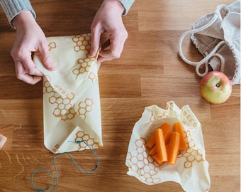 May include: A person is wrapping food in beeswax food wrap. The wrap has a honeycomb pattern and the text "Bees Wrap" is printed on it. The wrap is being used to wrap a piece of fruit.