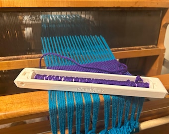 May include: A white plastic weaving tool with the text "priestlyavy" printed on it. The tool is being used to weave teal and purple yarn on a wooden loom.