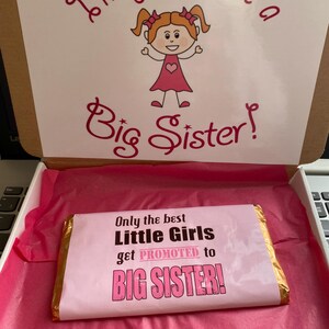 Könnte beinhalten: Eine rosa und goldene Schokoladentafel mit dem Text "Only the best Little Girls get PROMOTED to BIG SISTER!" Die Schokoladentafel befindet sich in einer rosa Geschenkbox mit einer weißen Karte, auf der "I'm going to be a Big Sister!" steht, und einem Comic-Mädchen mit Zöpfen.