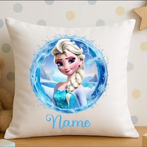 Op de afbeelding: Vierkantig wit kussen met een cartoonafbeelding van een vrouw met blond haar, gekleed in een blauwe jurk, omringd door een blauw waterontwerp. Het woord "Name" staat in lichtblauw script onderaan. Een houten ster en een teddybeer staan op de achtergrond.