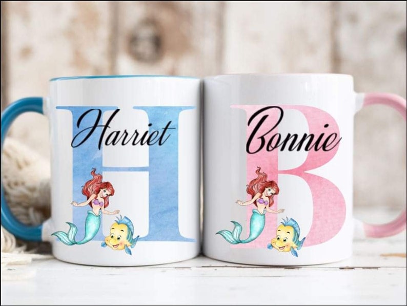 Może przedstawiać: Dwa białe ceramiczne kubki z niebieskimi i r&oacute;żowymi wzorami akwarelowymi. Kubki przedstawiają litery "H" i "B" z nazwami "Harriet" i "Bonnie" napisanymi powyżej. Każdy kubek ma ilustrację syreny i ryby.