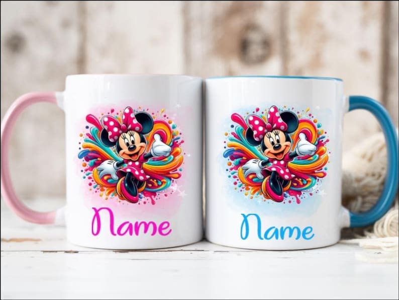 Op de afbeelding: Twee witte keramische mokken met roze en blauwe handvatten. Beide mokken hebben een kleurrijk ontwerp met een cartoonafbeelding van Minnie Mouse. De tekst "Name" is in roze en blauw op elke mok gedrukt.