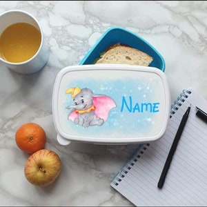 Könnte beinhalten: Eine weiße Brotdose mit blauem Deckel, verziert mit einer Illustration von Dumbo dem Elefanten und dem Wort "Name". Eine Tasse Orangensaft, ein Apfel und eine Mandarine liegen auf der Marmoroberfläche. Die Brotdose enthält ein Sandwich.