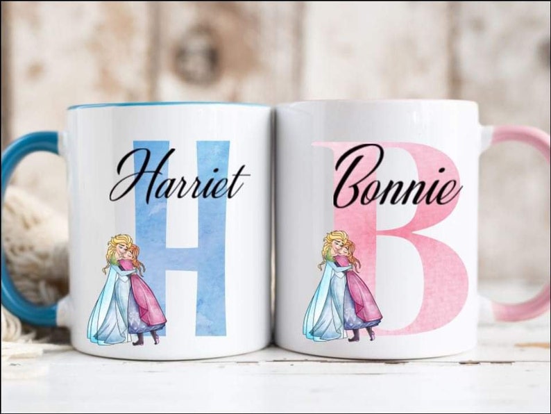 May include: Two white ceramic mugs with blue and pink handles. The mugs feature a watercolor design of the characters Elsa and Anna from the movie Frozen. The mugs are personalized with the names "Harriet" and "Bonnie" and their corresponding initial letters.