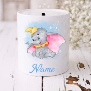 Puede incluir: Huchita de cerámica blanca con una imagen de dibujos animados de Dumbo el elefante con detalles azules, rosas y amarillos. El texto "Name" está escrito debajo de la imagen.