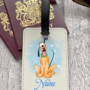 May include: White leather luggage tag with a cartoon image of Pluto the dog. Pluto is sitting with his tongue sticking out and a blue background with white stars. The text "Name" is written in blue at the bottom of the tag.