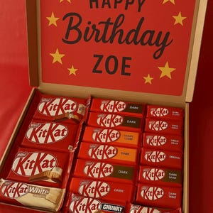 May include: A gift box filled with Kit Kat chocolate bars. The box lid is red with the words "HAPPY Birthday ZOE" and yellow star accents. Various Kit Kat flavors are visible.