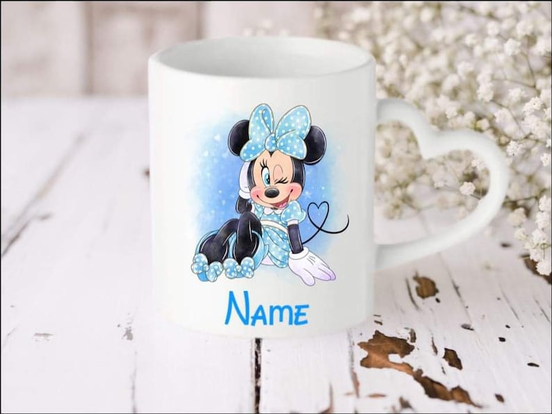 May include: White ceramic mug with a heart-shaped handle. The mug features a cartoon image of Minnie Mouse wearing a blue and white polka dot dress and a blue bow. The text "NAME" is printed below the image.