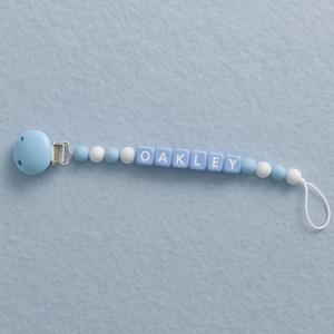 May include: A baby blue pacifier clip. The name "OAKLEY" is spelled out in white letters on blue square beads. The clip has a blue pacifier shield and a white loop at the end.