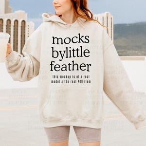 Puede incluir: Una sudadera con capucha beige con el texto "mocks bylittle feather" y el texto "this mockup is of a real model & the real POD item" impreso en la parte delantera.