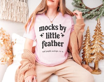 May include: A light pink short-sleeve t-shirt featuring the black text "mocks by little feather" with small star icons and the smaller text "real people mockups, no ai" printed on the front, worn with a tan knit cardigan and tan pants.