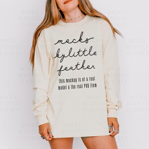May include: A cream coloured long-sleeved shirt with the text "mocks bylittle feather" and "this mockup is of a real model & the real POD item" printed on the front.