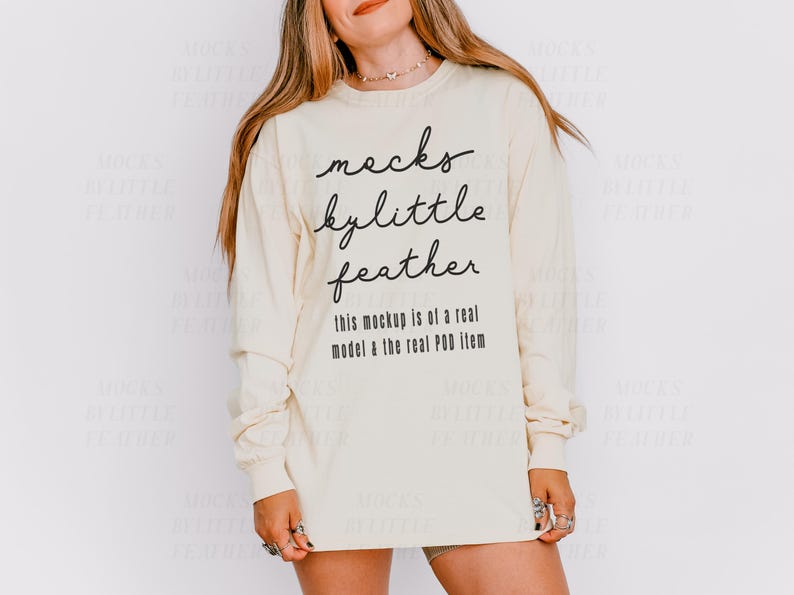 May include: A cream coloured long-sleeved shirt with the text "mocks bylittle feather" and "this mockup is of a real model & the real POD item" printed on the front.