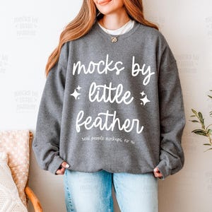 May include: A dark gray crewneck sweatshirt featuring the white text "mocks by little feather" in a script and sans-serif font combination, accented by two small four-pointed star icons, with the additional text "real people mockups, no ai".