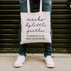Op de afbeelding: Een crèmekleurige canvas draagtas met twee schouderbanden, voorzien van de zwarte cursieve tekst "mocks by little feather" en de bloktekst "this mockup is of a real model & the real POD item" gedrukt op de voorzijde van de tas.