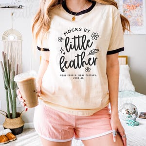 Next Level 3604 unisex ringer t-shirt mockup in natural with black trim, modeled indoors with a casual, boho vibe. The female model is styled in pink lounge shorts and accessorized with silver rings and layered gold jewelry, standing in a bright bedroom with cactus decor, a disco ball, and retro accents. Perfect for showcasing custom print-on-demand designs for Etsy listings, loungewear branding, and vintage-inspired apparel shops.
