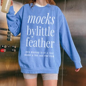Könnte beinhalten: Ein blaues langärmeliges Sweatshirt mit Rundhalsausschnitt und dem weißen Text "mocks bylittle feather" sowie "this mockup is of a real model & the real POD item" auf der Vorderseite. Das Kleidungsstück hat gerippte Bündchen am Kragen, an den Ärmeln und am Saum.