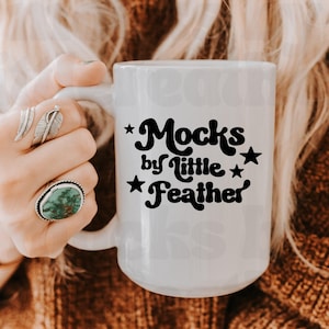 Op de afbeelding: Een witte keramische mok met zwarte tekst die Mocks by little Feather leest, geaccentueerd met kleine zwarte sterren. De mok heeft een glad, cilindrisch lichaam en een gebogen handvat aan de linkerkant voor een drankje.