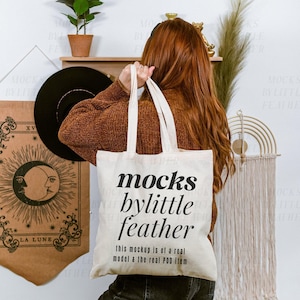 Peut inclure: Un sac fourre-tout en toile de couleur crème avec un texte imprimé en noir indiquant "mocks bylittle feather" suivi de "this mockup is of a real model & the real POD item" en plus petite police, avec deux longues anses.