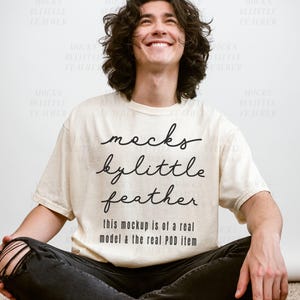 May include: A cream-colored short-sleeve t-shirt featuring black text that reads MOCKS BY LITTLE FEATHER and this mockup is of a real model & the real POD item. The shirt is worn with black distressed jeans and black sneakers.