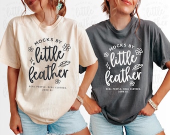 May include: Two short-sleeve crewneck t-shirts, one cream and one dark gray, each featuring the black or white text 'MOCKS BY little feather' with small flower, star, and leaf icons, plus the text 'REAL PEOPLE, REAL CLOTHES, ZERO AI.'