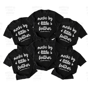 May include: A collection of six black short-sleeved t-shirts arranged in a cluster. Each shirt features white text that reads "mocks by little feather" with small star icons and smaller text below that reads "real people mockups, no ai".