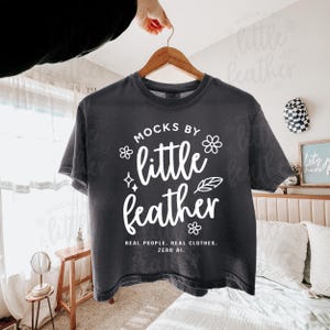 Comfort Colors 3023CL cropped tee mockup in Pepper, displayed on a wooden hanger held in a softly lit, boho-inspired bedroom. This simple and trendy Comfort Colors mockup by MocksByLittleFeather features the C3023CL shirt with real fabric texture and no AI—perfect for Etsy listings, print-on-demand shops, and aesthetic apparel branding.