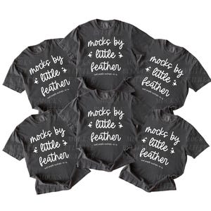 May include: A collection of six dark gray short-sleeve t-shirts arranged in a cluster. Each shirt features white text that reads "mocks by little feather" in a script font, accompanied by small four-pointed star icons on either side of the word little.