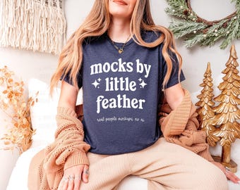 May include: A dark navy blue short-sleeved t-shirt featuring white text that reads mocks by little feather with two small star icons, and smaller text below that reads real people mockups no ai, worn with a tan knit cardigan.