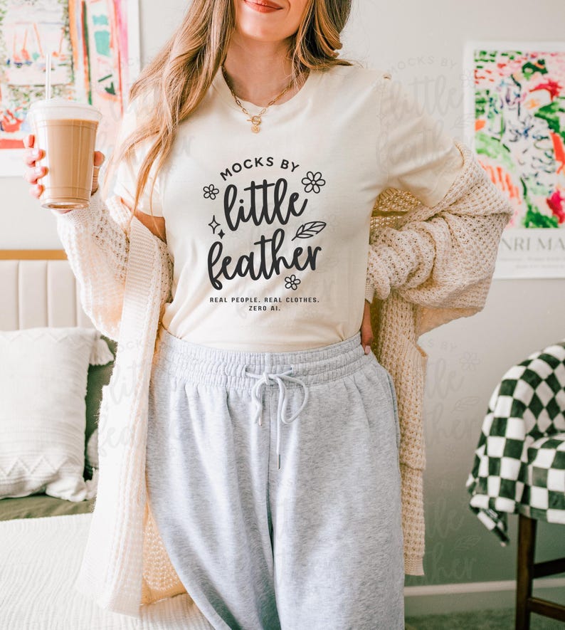 Woman wearing natural Bella Canvas 3001 t-shirt with 'Mocks by Little Feather' logo design, styled with cozy knit cardigan and gray sweatpants in a modern bedroom setting — trendy real people mockup for Etsy sellers and print-on-demand apparel