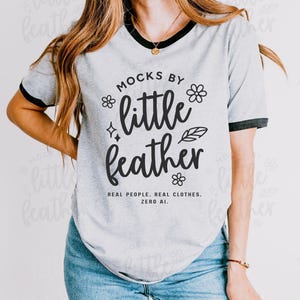 Next Level 3604 unisex ringer t-shirt mockup in heather grey with black trim, featuring a smiling female model styled in light wash jeans and layered gold jewelry. Set against a minimalist studio background, this lifestyle mockup is ideal for showcasing custom t-shirt designs for Etsy, print-on-demand shops, retro-inspired apparel, and everyday casual fashion branding.