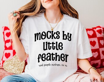 May include: A white short-sleeve t-shirt featuring the black text "mocks by little feather" and "real people mockups, no ai" printed on the front. The shirt has a crew neckline and is made of a plain white fabric.