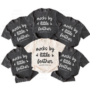May include: A collection of five short-sleeved t-shirts arranged in a group. Four shirts are dark charcoal gray with white text reading "mocks by little feather" and small star icons. One central shirt is cream with black text and star icons.