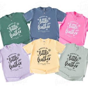 May include: Six short-sleeve crewneck t-shirts in green, blue, pink, lavender, yellow, and light gray. Each shirt features the white or black printed text MOCKS BY little feather, accompanied by small flower, star, and leaf graphic icons.