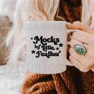 Op de afbeelding: Een witte keramische mok met zwarte tekst die Mocks by little Feather leest, geaccentueerd met kleine zwarte sterren. De mok heeft een glad, cilindrisch lichaam met een gebogen handvat aan de rechterkant, vastgehouden voor het ontwerp.