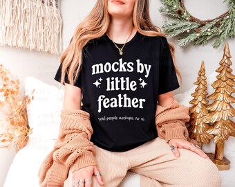 May include: A black short-sleeve t-shirt featuring white text that reads "mocks by little feather" with small star icons and the smaller text "real people mockups; no ai" underneath, worn with a tan knit cardigan.