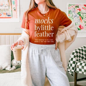May include: A woman wearing a rust-colored t-shirt with the text "mocks bylittle feather" and the words "this mockup is of a real model & the real POD item" printed on the shirt. She is wearing gray sweatpants and a cream cardigan.