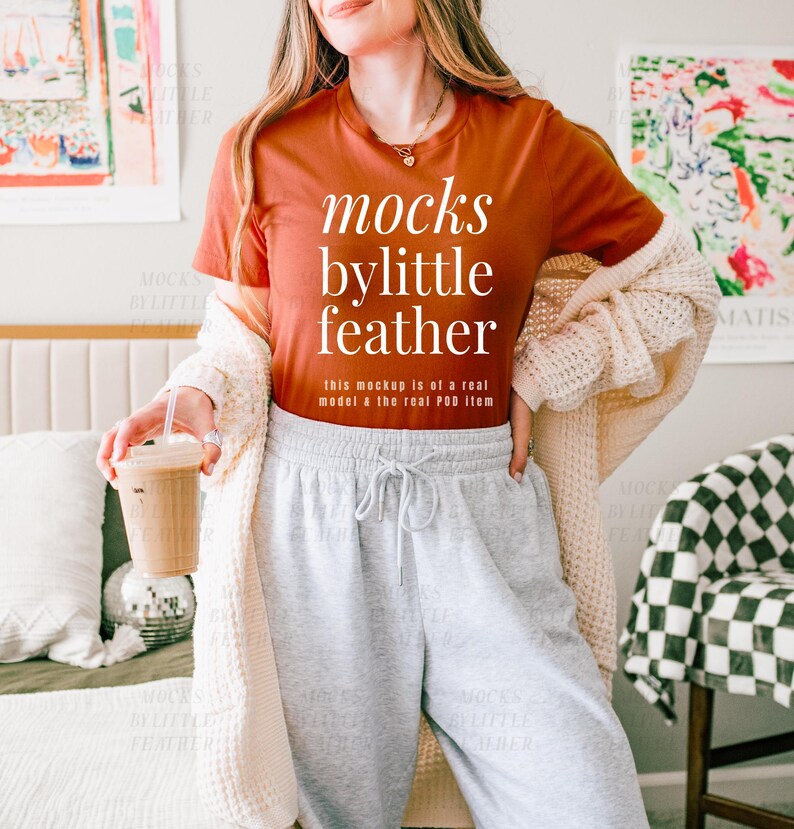 May include: A woman wearing a rust-colored t-shirt with the text "mocks bylittle feather" and the words "this mockup is of a real model & the real POD item" printed on the shirt. She is wearing gray sweatpants and a cream cardigan.