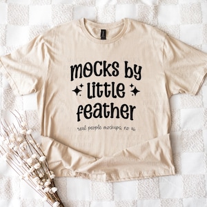 Peut inclure: Un t-shirt beige à manches courtes avec le texte noir "mocks by little feather" accompagné d'étoiles, et une ligne plus petite indiquant "real people mockups, no ai". Le vêtement présente un col rond et une coupe classique.