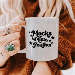 Op de afbeelding: Een witte keramische mok met zwarte tekst die Mocks by little Feather leest, geaccentueerd met kleine zwarte sterren. De mok heeft een glad, cilindrisch lichaam met een gebogen handvat aan de zijkant, met een strak ontwerp.