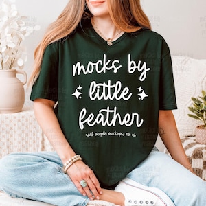 May include: A dark green short-sleeve t-shirt featuring white text that reads "mocks by little feather" with two small star icons. The shirt has a crew neckline and a relaxed fit, with additional small text below reading "real people mockups, no ai".