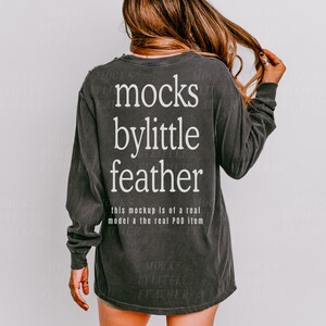 May include: A dark grey long-sleeved shirt with the text "mocks bylittle feather" printed on the back. The text is in white and is centred on the shirt. The shirt is a relaxed fit and has a distressed look.