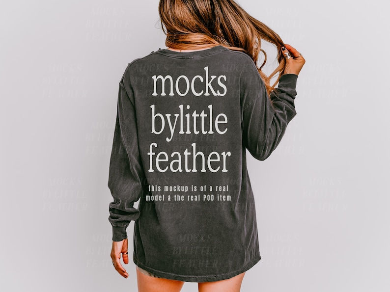 May include: A dark grey long-sleeved shirt with the text "mocks bylittle feather" printed on the back. The text is in white and is centred on the shirt. The shirt is a relaxed fit and has a distressed look.