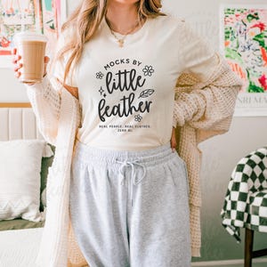 Woman wearing natural Bella Canvas 3001 t-shirt with 'Mocks by Little Feather' logo design, styled with cozy knit cardigan and gray sweatpants in a modern bedroom setting — trendy real people mockup for Etsy sellers and print-on-demand apparel