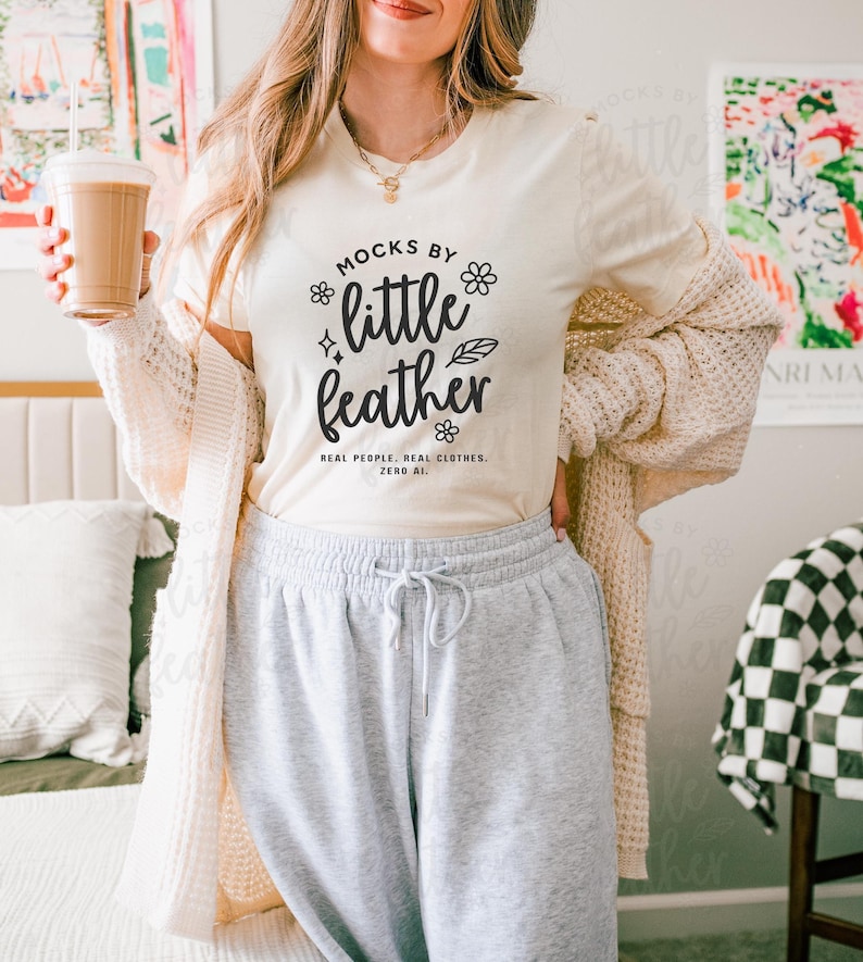 Woman wearing natural Bella Canvas 3001 t-shirt with 'Mocks by Little Feather' logo design, styled with cozy knit cardigan and gray sweatpants in a modern bedroom setting — trendy real people mockup for Etsy sellers and print-on-demand apparel
