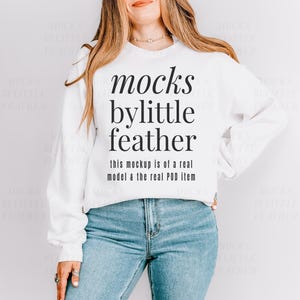 May include: A white long-sleeve crewneck sweatshirt featuring black text that reads: mocks bylittle feather, this mockup is of a real model & the real POD item. The sweatshirt is paired with blue denim jeans, showcasing a casual and clean aesthetic.