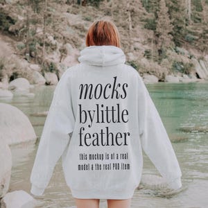 Puede incluir: Una sudadera con capucha blanca con el texto "mocks by little feather this mockup is of a real model & the real POD item" impreso en la parte posterior.